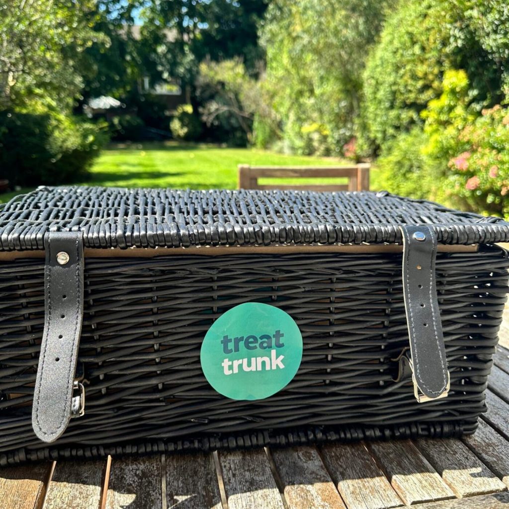 The Treat Trunk Signature Hamper: a black wicker corporate Christmas hamper with leather straps and buckles, on a wooden garden table